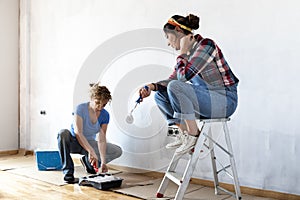 Two women renovating a house. Loading roller in paint tray. Sitting on painter ladder. Painting house walls white