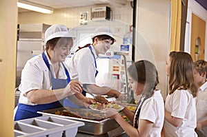 Two women serving food to a girl in a school cafeteria queue