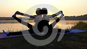 Two Women Sit Back-To-Back on Mat And Raise One Leg Each at Sunset