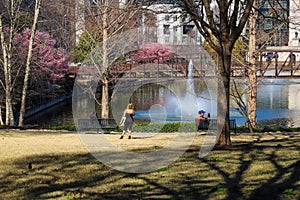 Two women sitting on black metal benches and one woman standing near a green lake in the park surrounded by pink trees