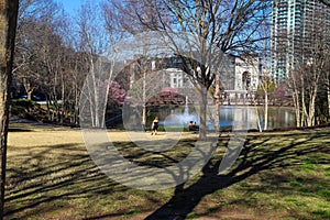 Two women sitting on black metal benches and one woman standing near a green lake in the park surrounded by pink trees