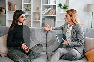 Two women is sitting on the sofa and talking. Casual and formal clothes