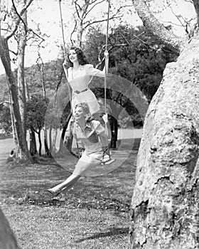 Two women sitting and standing on a swing photo