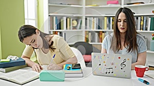 Two women sitting on table studying using laptop writing notes at library university