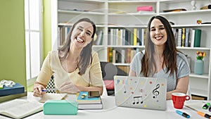 Two women sitting on table studying using laptop writing notes at library university