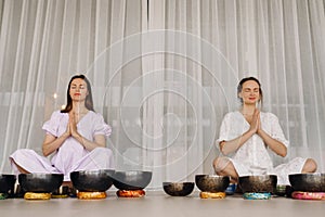 Two women are sitting with Tibetan bowls in the lotus position before a yoga class in the gym