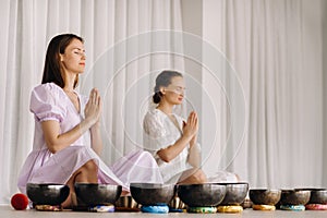 Two women are sitting with Tibetan bowls in the lotus position before a yoga class in the gym