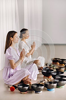 Two women are sitting with Tibetan bowls in the lotus position before a yoga class in the gym