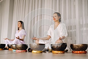 Two women are sitting with Tibetan bowls in the lotus position before a yoga class in the gym