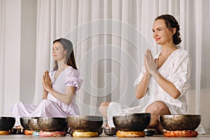 Two women are sitting with Tibetan bowls in the lotus position before a yoga class in the gym
