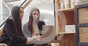 Two women sitting and working in a modern and industrial style cafe. Two multiethnic colleagues working on laptop and