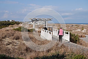 Two women stroll across a bridge in the dunes to a wooden archway to a roadway