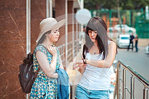 Two women in summer clothes are standing on the street, and look at the smart watch on their hand. Outdoor. Concept of modern