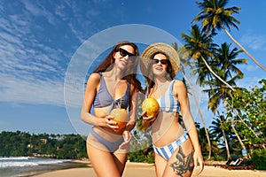 Two women in swimsuits hold coconuts, enjoying sun on a tropical beach with palm trees. Friends share a laugh, enjoy