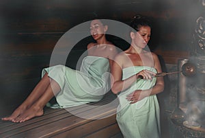 Two women in a traditional steam sauna banya pours water on hot stones on the stove