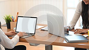 Two women are using a white blank screen computer laptop on the wooden working desk.