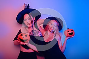 Two women witches hold pumpkins in their hands and dance. Women in neon lights are celebrating a Halloween party. Models smile and