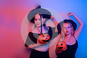 Two women witches hold pumpkins in their hands and dance. Women in neon lights are celebrating a Halloween party. Models smile and