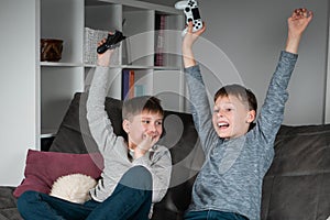 Two wonderful positive teenage boys sitting on sofa at home, holding gaming controller, playing videogames, celebrating.