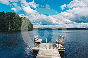 Two wooden chairs bench on a wood pier overlooking a blue lake water with green forest and cloud sky