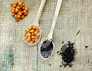 Two wooden spoons with vitaminic healthy sea buckthorn berries and black tea over sackcloth background. cooking,