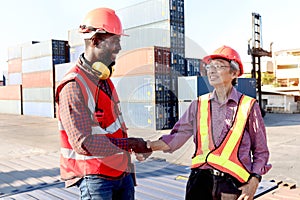 Two worker wear safety helmet and safety red vest shaking hands at logistic shipping cargo containers yard. African engineer man