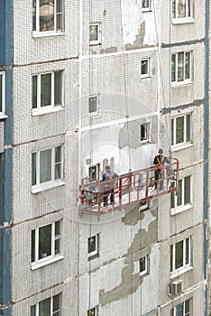 Two workers in construction cradle on metal cables repairing vertical iwall surface on block of flats house