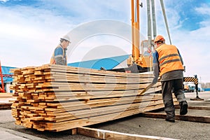 Two workers in helmets and construction vests unload wooden planks. Slingers stack lumber outside in open air.