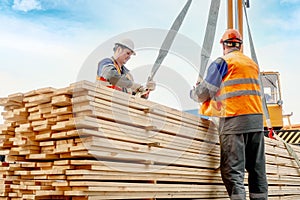 Two workers in helmets and construction vests unload wooden planks. Slingers stack lumber outside in open air.