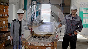 Two workers are holding a hanging box with finished metal details, walking in a space of workshop