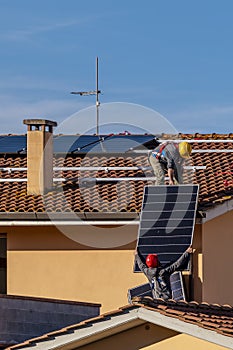 Two workers pass solar panels to each other to be mounted on top of a red tiled roof to install a photovoltaic system