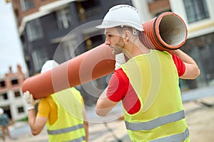 Two workers in protective helmets carrying a corrugated drain pipe