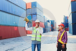 Two workers with safety vest and helmet working at logistic shipping cargo containers yard, the engineer boss holding megaphone