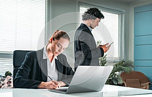 Two working concentrated business people, one is sitting at the desk in front of laptop, second is using smartphone at office