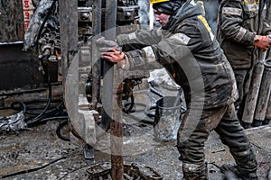 Two working drillers in a uniform, in a helmet and goggles, install drill pipes after lifting them from an oil well
