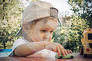 A two year old boy rolls a small toy car on a table outdoors. A child plays with small cars on a summer day.