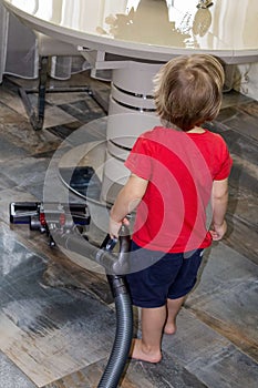 A two-year-old boy vacuums the floor in a living room, the concept of child labor and helping his mother/