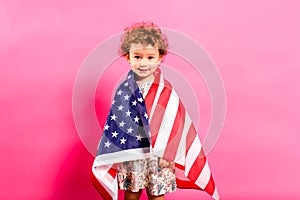 Two year old girl posing sympathetic with the American flag on her shoulders, isolated on a pink background