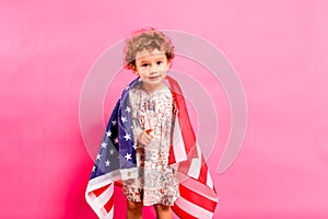 Two year old girl posing sympathetic with the American flag on her shoulders, isolated on a pink background