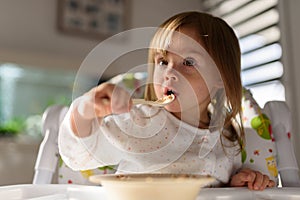 Two years old eats brakefast by herself with a spoon.