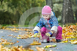 Two years old girl stretching her hand to metal thermos flask cup hunkering at autumn foliage covering backdrop