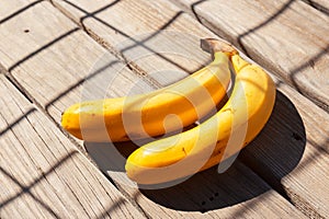 Two yellow bananas on the wooden surface in chain fence shadow. Fresh organic fruit on sunny day. Close up. Selective focus.