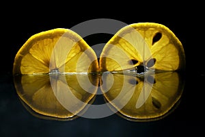 Two yellow fresh lemon cuts slices backlit isolated on black background with reflection close-up