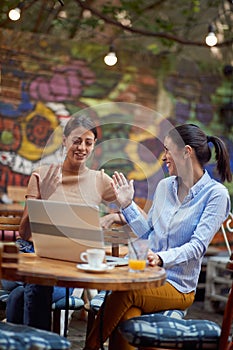 Two young adult women sitting in outdoor cafe, smiling, with palms  in the air preparing for high five