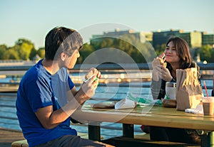 Two young adults eating hamburgers outdoors by lake, urban setting