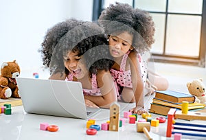 Two young African girls play with notebook computer among toys, doll and book in front of glass window