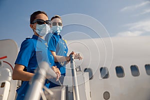 Two young air stewardesses in blue uniform, sunglasses and protective face masks looking at camera, standing on airstair