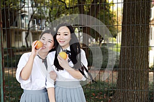 Two young Asian Chinese pretty girls wear student suit in school best friends smile laugh smell orange fruit in nature