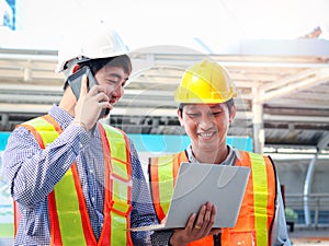 Two young Asian male engineer workers wearing safety vest and helmet, looking at laptop computor together, discussing about