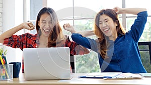 Two young asian women using laptop computer and arms up with smiling face while working at home office desk, success in business